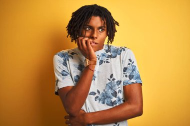 Izole sarı arka plan üzerinde yaz çiçek t-shirt giyen dreadlocks ile Afro adam stresli ve ağız tırnak ısırma elleri ile sinir görünümlü. Anksiyete sorunu.