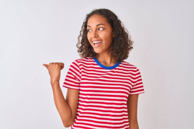 Kırmızı çizgili tişört giyen genç Brezilyalı bir kadın izole edilmiş beyaz arka planda gülümsüyor ve başparmağıyla yan tarafa bakıyor..