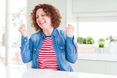 Kıvırcık saçlı, kot ceket giyen orta yaşlı bir kadın kolları havada ve gözleri kapalı bir şekilde başarısını kutluyor ve şaşırıyor. Kazanan konsept.
