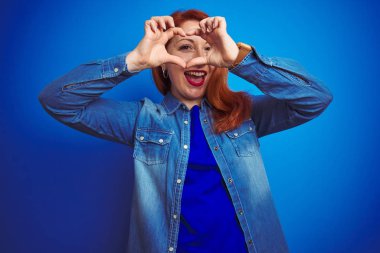 Genç, güzel, kızıl saçlı, kot gömlek giyen, mavi izole edilmiş arka planda duran el ve parmaklarıyla kalp şeklinde gülümseyen bir kadın.