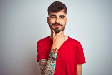 Kırmızı t-shirt giyen dövme giyen genç adam izole beyaz arka plan üzerinde duran çapraz kolları ve çene üzerinde kaldırdı el ile gülümseme ile kameraya emin bakıyor. Olumlu düşünme.