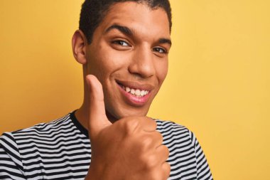 Genç, yakışıklı, denizci çizgili tişört giyen, izole edilmiş sarı arka planda mutlu, gülümseyen, işaret veren, parmaklarıyla başparmağını kaldıran, mükemmel bir işaret.