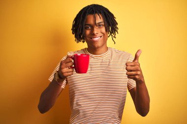 Dreadlocks ile Afro amerikan adam izole sarı arka plan üzerinde bir fincan kahve içme büyük bir gülümseme ok işareti yapıyor mutlu, parmakları ile başparmak, mükemmel işareti