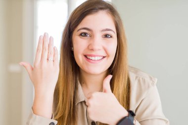 Beautiful young woman showing engagement ring on hand happy with big smile doing ok sign, thumb up with fingers, excellent sign clipart