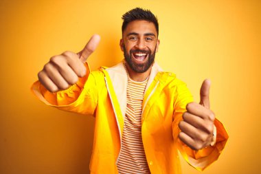 İzole sarı arka plan üzerinde duran yağmurluk giyen genç hintli adam el iyle olumlu jest yaptığını onaylayarak, gülümseyerek ve başarı için mutlu başparmak. Kazanan jest.