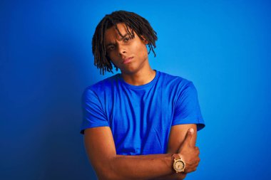 Izole mavi arka plan şüpheci ve sinir üzerinde duran t-shirt giyen dreadlocks giyen Afro Amerikan adam, çapraz kolları ile yüzünde onaylamayan ifade. Negatif kişi.
