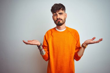 Dövme giyen genç adam izole beyaz arka plan üzerinde duran turuncu kazak habersiz ve kolları ve elleri ile karışık ifade kaldırdı. Şüphe kavramı.