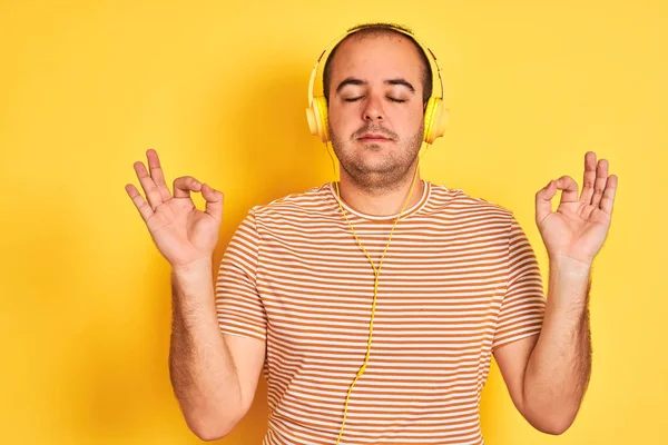 孤立した黄色の背景の上に立ってヘッドフォンを使用して音楽を聞いて若い男はリラックスし 指で瞑想のジェスチャーを行う目を閉じて笑顔 ヨガのコンセプト — ストック写真