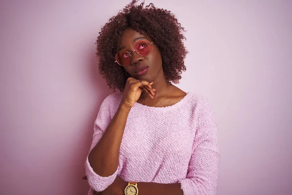 Young african afro woman wearing sweater and sunglasses over isolated pink background with hand on chin thinking about question, pensive expression. Smiling with thoughtful face. Doubt concept.