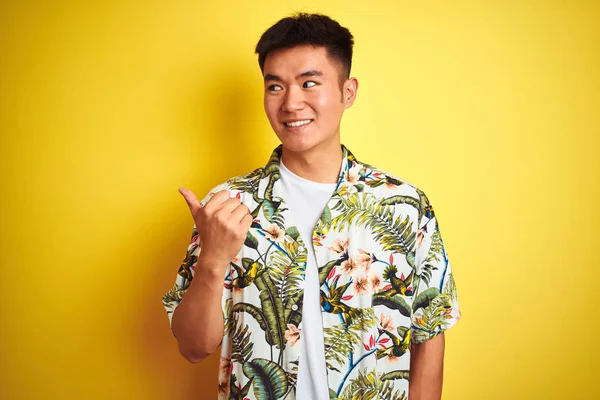 Asian chinese man on holiday wearing summer shirt over isolated yellow background smiling with happy face looking and pointing to the side with thumb up.