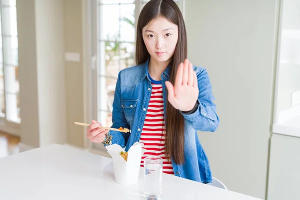 Beautiful Asian woman eating asian rice in delivery box with open hand doing stop sign with serious and confident expression, defense gesture