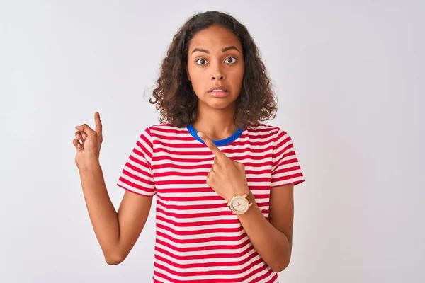 Young brazilian woman wearing red striped t-shirt standing over isolated white background Pointing aside worried and nervous with both hands, concerned and surprised expression