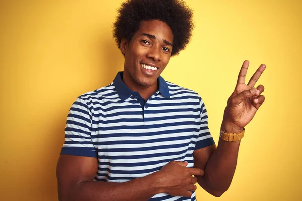 American man with afro hair wearing navy striped polo standing over isolated yellow background smiling with happy face winking at the camera doing victory sign. Number two.