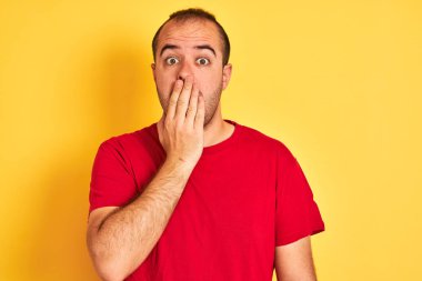 Kırmızı günlük tişört giyen genç adam izole edilmiş sarı arka planda duruyor. Ağzı kapalı, elleri utançla dolu, hata yüzünden şoka girmiş, korku ifadesi, sessizlikten korkmak, gizli bir konsept.