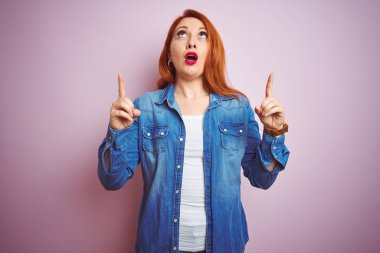 Izole pembe arka plan üzerinde duran denim gömlek giyen güzel kızıl saçlı kadın Youg şaşırmış ve baktı ve parmakları ile işaret ve kolları kaldırdı şaşırttı.