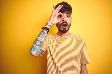 Dövmesi olan genç adam çizgili t-shirt giyen izole sarı arka plan üzerinde duran ok jest yaparken şaşırmış yüz ile şok, parmaklardan bakarak göz. İnanmayan ifade.