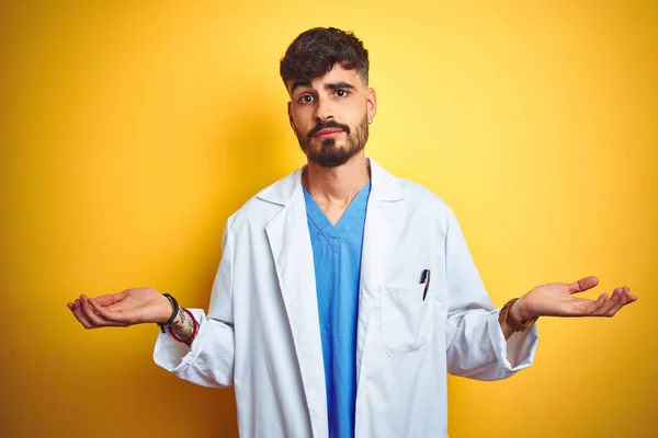 Young doctor man with tattoo standing over isolated yellow background clueless and confused expression with arms and hands raised. Doubt concept.