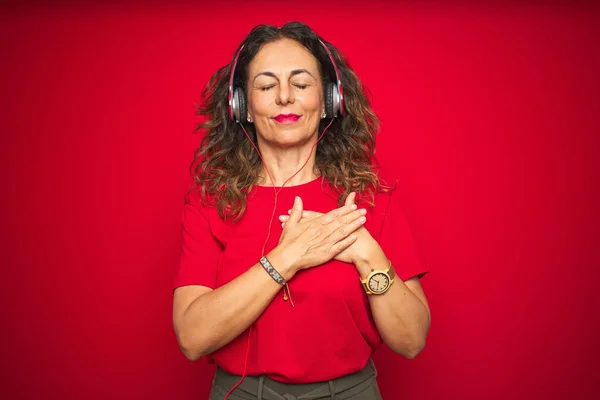 ヘッドフォンを装着した中年の先輩女性が 目を閉じて感謝のジェスチャーをして胸に微笑む赤い孤立した背景の上で音楽を聴いている 健康の概念 — ストック写真
