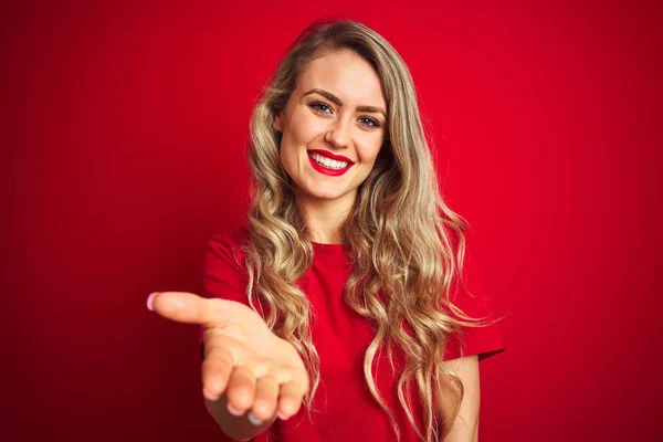 赤い孤立した背景の上に立つ基本的なTシャツを着た若い美しい女性は 挨拶と歓迎として握手を提供する友好的な笑顔 ビジネスの成功 — ストック写真