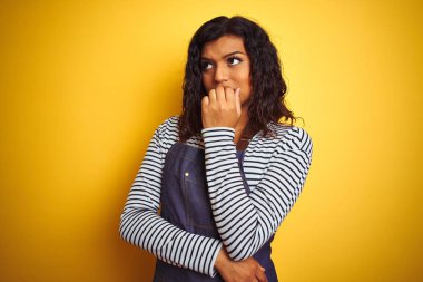 Genç transseksüel transseksüel şef kadın izole sarı arka plan üzerinde önlük giyen stresli ve ağız tırnak ısırma elleri ile sinirli görünümlü. Anksiyete sorunu.