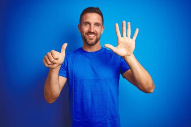 Genç yakışıklı adam mavi izole arka plan üzerinde rahat t-shirt giyen gösteren ve emin ve mutlu gülümserken parmakları sayısı altı ile işaret.