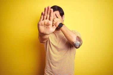 Dövme ile genç yakışıklı adam izole sarı arka plan üzerinde çizgili t-shirt giyen elleri ile gözleri kaplayan ve üzgün ve korku ifadesi ile stop jest yapıyor. Utanmış ve olumsuz kavram.