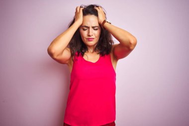 Pembe izole edilmiş arka planda duran tişört giyen genç ve güzel bir kadın. Baş ağrısı, acı ve migren yüzünden çaresiz ve stresli. Ellerini başının üstüne koy..