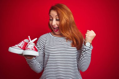 Güzel, kızıl saçlı, spor ayakkabılarını kırmızı arka planda tutan genç bir kadın gururla bağırıyor, zaferi ve başarıyı kutluyor çok heyecanlı, coşkulu bir duygu.