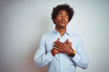 Afro saçlı genç afro-amerikan adam izole beyaz arka plan üzerinde kapalı gözleri ve yüzünde minnettar jest ile göğüs üzerinde gülümseyerek zarif gömlek giyiyor. Sağlık kavramı.