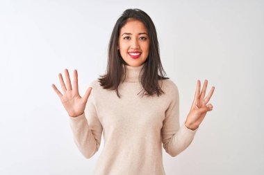 Balıkçı yaka kazak giyen güzel Çinli kadın izole edilmiş beyaz arka plan üzerinde duruyor ve parmaklarını sekiz numaralı parmaklarıyla gösterirken kendinden emin ve mutlu bir şekilde gülümsüyor..