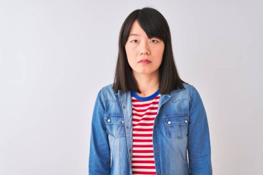 Çinli kadın yüzünde ciddi ifade ile izole beyaz arka plan üzerinde denim gömlek ve kırmızı çizgili t-shirt giyiyor. Kameraya basit ve doğal bir bakış.