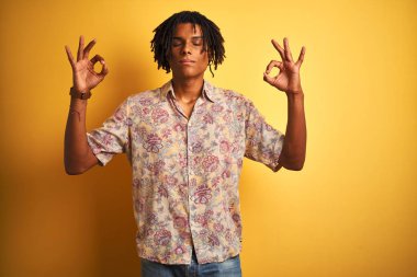 Izole sarı arka plan üzerinde çiçek gömlek giyen tatilde dreadlocks ile Afro adam rahatlayın ve parmakları ile meditasyon jest yapıyor kapalı gözleri ile gülümseyerek. Yoga konsepti.