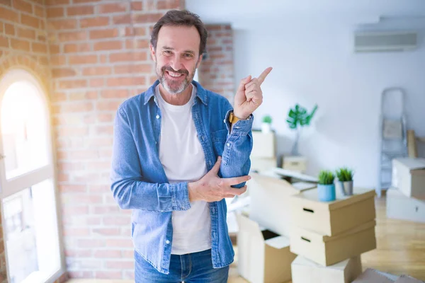 Middle age senior man moving to a new house packing cardboard boxes with a big smile on face, pointing with hand and finger to the side looking at the camera.