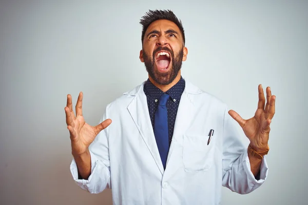 Young indian doctor man standing over isolated white background crazy and mad shouting and yelling with aggressive expression and arms raised. Frustration concept.