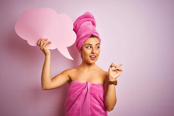 Beautiful woman wearing towel holding cloud speech bubble over isolated pink background very happy pointing with hand and finger to the side