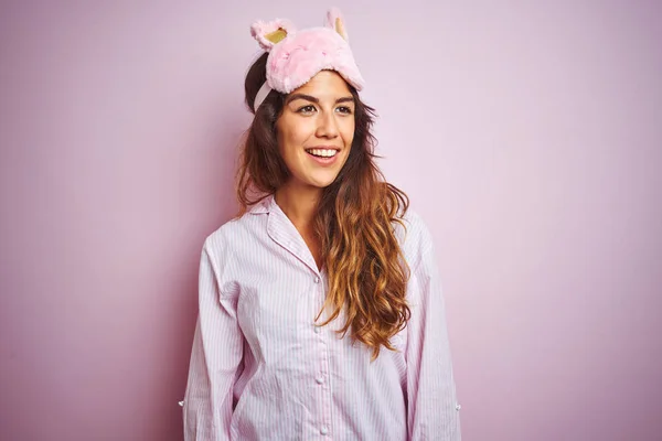 Young woman wearing pajama and sleep mask standing over pink isolated background looking away to side with smile on face, natural expression. Laughing confident.