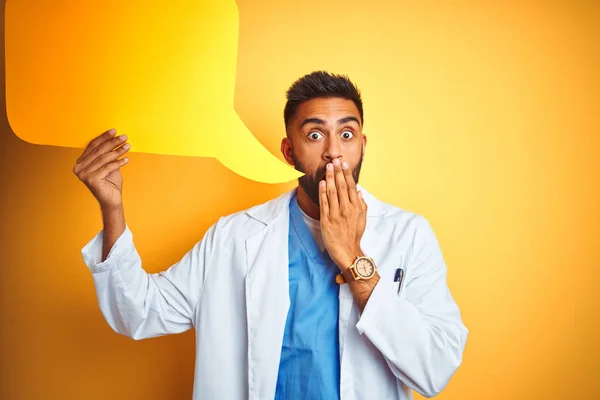 Young indian doctor man holding speech bubble standing over isolated yellow background cover mouth with hand shocked with shame for mistake, expression of fear, scared in silence, secret concept