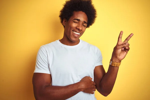 American man with afro hair wearing white t-shirt standing over isolated yellow background smiling with happy face winking at the camera doing victory sign. Number two.