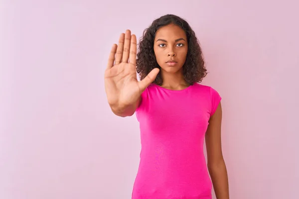 隔離されたピンクの背景の上に立つTシャツを着た若いブラジル人女性は 手の手のひらで歌うのをやめます 顔に負と深刻なジェスチャーで警告式 — ストック写真