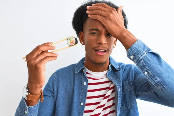 Young african american man eating sushi using chopsticks over isolated white background stressed with hand on head, shocked with shame and surprise face, angry and frustrated. Fear and upset for mistake.