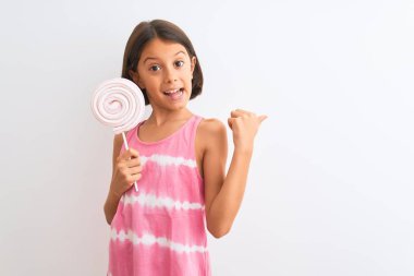 Güzel çocuk kız izole beyaz arka plan üzerinde duran tatlı lolipop yeme işaret ve mutlu yüz gülümseyerek yan başparmak ile gösteren