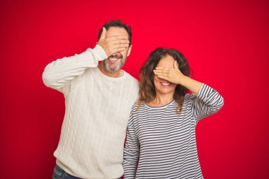 Beautiful middle age couple wearing winter sweater over isolated red background smiling and laughing with hand on face covering eyes for surprise. Blind concept.