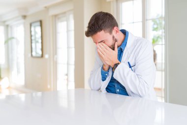 Handsome doctor man wearing medical coat at the clinic with sad expression covering face with hands while crying. Depression concept.