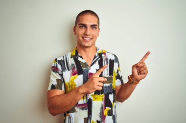 Beyaz tenli arka planda yaz tişörtü giyen yakışıklı genç adam gülümsüyor ve kameraya bakıyor iki eliyle ve parmaklarıyla kenarı işaret ediyor..