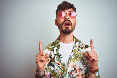 Izole beyaz arka plan üzerinde yazlık gömlek güneş gözlüğü giyen tatilde dövme ile Adam şaşırttı ve yukarı bakarak ve parmakları ile işaret ve kolları kaldırdı şaşırttı.