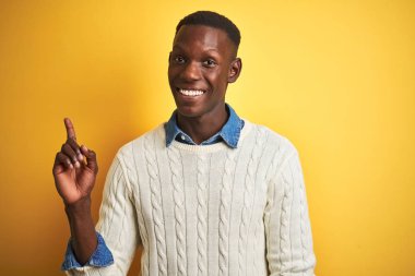 Denim gömlek ve beyaz kazak giyen afro-amerikan adam yüzünde büyük bir gülümseme ile izole sarı arka plan üzerinde, kameraya bakarak yan el parmağı ile işaret.