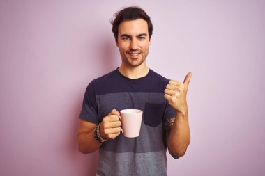 Izole pembe arka plan üzerinde kahve dövme içme fincan ile genç yakışıklı adam ok işareti yapıyor büyük bir gülümseme ile mutlu, parmakları ile başparmak, mükemmel işareti