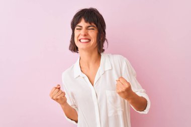 Izole pembe arka plan üzerinde duran beyaz gömlek giyen genç güzel kadın çok mutlu ve heyecanlı kolları kaldırdı ile kazanan jest yapıyor, gülümseyerek ve başarı için çığlık. Kutlama konsepti.