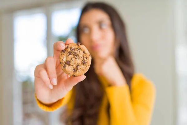 Young woman eating chocolate chips cookies at home serious face thinking about question, very confused idea