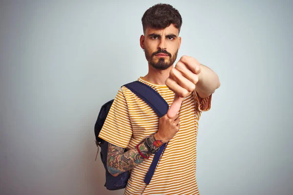 Young student man with tattoo wearing backpack standing over isolated white background with angry face, negative sign showing dislike with thumbs down, rejection concept